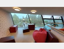 Moderner Innenraum mit großen, schrägen Glasfenstern, die Ausblick auf grüne Bäume bieten. Der Raum ist mit roten Sitzmöbeln, einem Schreibtisch, einem Stuhl sowie einer roten Sitzsack-Liege ausgestattet und zeigt eine geschwungene Glasbrüstung.