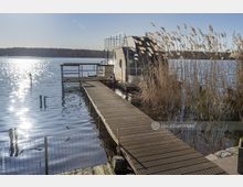 Hausboot an einem See, umgeben von Schilfgras und verbunden mit einem hölzernen Steg unter klarem Himmel. Im Hintergrund erstreckt sich bewaldetes Ufer und die Wasseroberfläche reflektiert Sonnenlicht.