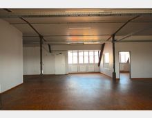 Ein leerer, moderner Raum mit Holzfußboden, weißen Wänden und einer Decke mit offenen Metallträgern. An der Stirnseite befinden sich Fenster, Heizkörper und eine weiße Tür, daneben ein Zugang zu einem weiteren Raum.