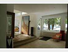 Heller Innenraum mit großen Fenstern, durch die Bäume und parkende Autos sichtbar sind. Im Vordergrund ein gefliester Boden, ein orangefarbener Stuhl, und ein gerahmtes Bild einer Giraffe, das an einer Wand neben einer kleinen Treppe lehnt.