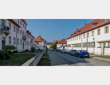 Straße mit gepflastertem Gehweg und parkenden Autos, flankiert von weißen Wohnhäusern mit roten Ziegeldächern und Fensterläden. Im Hintergrund sind Bäume und ein modernes Gebäude sichtbar.