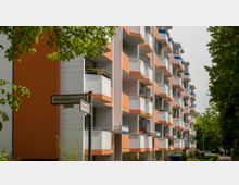 Mehrstöckiges Wohngebäude mit Balkonen in Weiß und Orange, umgeben von Bäumen und Grünflächen; im Vordergrund ein Straßenschild mit der Aufschrift „Bärensteinstraße“.