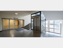 Foyerbereich eines Gebäudes mit Fliesenboden, einem Aufzug mit Glastüren, einer Treppe mit Metallgeländer und großen Fenstern mit satiniertem Glas. Links befindet sich eine doppelflügelige Glastür mit der Aufschrift „Zu den Sälen“.