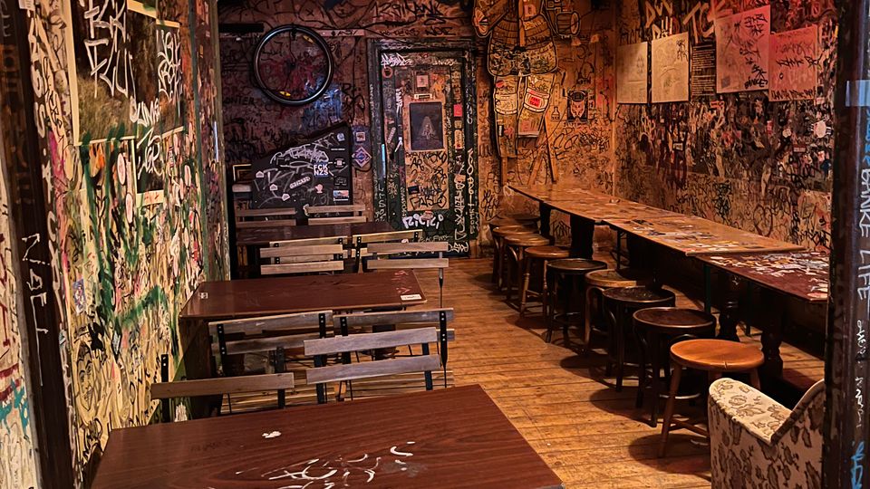 Schmaler Innenraum eines alternativ gestalteten Cafés oder Bars mit Holzdielenboden, langen Holztischen und Hockern entlang der rechten Wand sowie Bänken links. Die Wände und die Tür sind dicht mit Graffiti, Stickern und Zeichnungen bedeckt, beleuchtet von mehreren hängenden Lampenschirmen in warmen Farben.