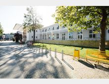 Öffentlicher Platz vor einem modernen Gebäude mit weißer Fassade, großen Fenstern und einem Vordach. Im Vordergrund befinden sich ein gepflasterter Weg, Fahrradständer, eine gelbe Bank und ein Mülleimer sowie Rasenflächen mit Schatten spendenden Bäumen.