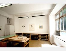 Heller Raum mit weiß gestrichenen Wänden, großen Fenstern auf der rechten Seite und einfachen Industriemöbeln aus Holzpaletten und Arbeitsflächen. An den Wänden hängen Bilder und Zeichnungen, während Leuchtstofflampen von der Decke hängen.