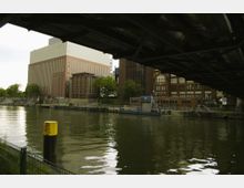 Blick auf industrielle Gebäude aus rotem und hellem Mauerwerk am Ufer eines Flusses, aufgenommen von unter einer Brücke. Im Vordergrund sind ein gelber Poller, ein Zaun und reflektierendes Wasser sichtbar.