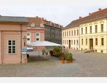 Pflastersteingasse in einer historischen Stadt mit pastellfarbenen Gebäuden. Links ein rosafarbenes Haus mit einem Hinweisschild "Restaurant Waage", rechts ein gelbes Gebäude mit verzierten Fassaden und roten Fensterrahmen, in der Mitte ein Sonnenschirm und Pflanzkübel.