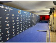 Ein langgestreckter Raum mit einem blauen Bodenbelag und einer Wand mit Prime-Video-Logos sowie Logos einer Show. Auf der rechten Seite stehen Lampenstative für Beleuchtung, und im Hintergrund ist eine rote Wand mit einer Tür sichtbar.