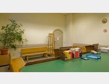 Ein einfacher Raum mit hellen Wänden, einer Holztür mit bogenförmigem Rahmen und einer hölzernen Sitzbank an der Wand. Im Vordergrund steht ein großer Tisch mit grüner Oberfläche, auf dem Bücher, Büroartikel und eine blaue Ablage liegen; im Hintergrund sind ein Kleiderständer und eine Zimmerpflanze zu sehen.