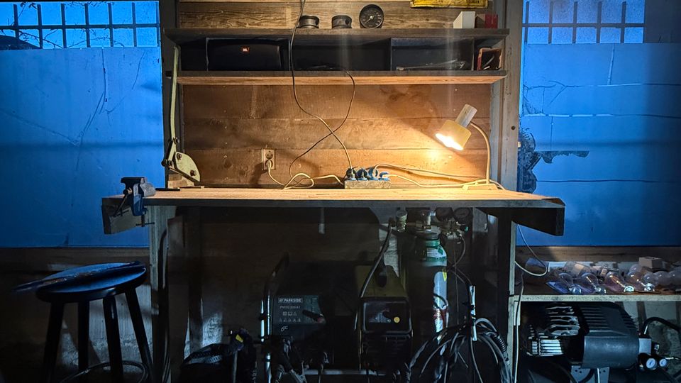 Werkstatt- oder Garageninnenraum mit hölzerner Werkbank und Regal, beleuchtet von einer hängenden Lampe und einer kleinen Arbeitsleuchte. Links und rechts befinden sich zwei Fenster; unter der Werkbank stehen Maschinen und Kabel, daneben ein großer Kompressor.