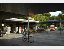 Ein kleiner gepflasterter Platz mit Überdachung und mehreren Geschäften, darunter eine "Fotothek". Im Vordergrund steht ein Fahrrad an einem Laternenpfahl, im Hintergrund parken ein Krankenwagen und ein grünes Polizeifahrzeug. Im Hintergrund sind Bäume zu sehen.