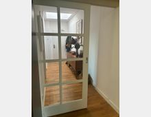 Ein Zuhauseingangsbereich mit Holzboden, einer weißen Schiebetür mit Glasfeldern und einer Anrichte an der Wand. Auf der Anrichte stehen eine Vase mit Blumen und andere Gegenstände, während darüber ein gerahmtes Bild hängt.