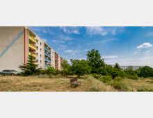 Mehrgeschossiges Wohngebäude mit bunten Balkonreihen links im Bild, umgeben von Bäumen und einer Wiese mit einer einfachen Holzbänken im Vordergrund. Im Hintergrund sind weitere Wohnblöcke sowie ein blauer Himmel mit Wolken zu sehen.
