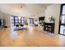 Großzügiges modernes Wohnzimmer mit offenem Grundriss, gestaltet mit hellem Holzboden, großen Fensterfronten und weißen Vorhängen. Der Raum umfasst einen Essbereich mit Tisch und Stühlen, eine Sitzecke mit Sofas und eine offene Küche mit schwarzen Schränken, dekoriert mit Pflanzen und kleinen Akzenten.