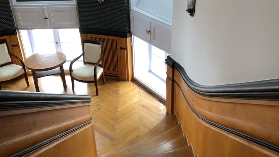 Blick von einer geschwungenen Holztreppe in ein helles Treppenhaus mit Fischgrätparkett, weißen Wandpaneelen und dunkler gemusterter Wandverkleidung. Große Rund- und Dachfenster sowie Wandlampen beleuchten eine kleine Sitzecke mit zwei Stühlen und einem runden Tisch.