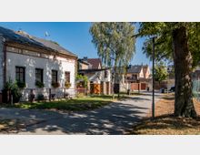 Wohnviertel mit gepflasterter Straße und Gehweg, links ein weißes, eingeschossiges Haus mit Blumen an den Fenstern, umgeben von einem Gartenzaun. Im Hintergrund weitere Wohnhäuser mit modernen Dächern sowie Bäume und ein einzelner Straßenlaternenmast.