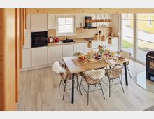 Moderne Wohnküche mit einer hellen Holzoptik, einem Esstisch aus Holz mit geflochtenen Stühlen, die mit Fellauflagen dekoriert sind. Im Hintergrund befinden sich eine minimalistische Küchenzeile mit eingebauten Schränken, ein großes Fenster, das Tageslicht hereinlässt, und ein Kamin auf der rechten Seite.