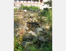 Kleiner Gartenwasserfall, der über eine mit Steinen und Felsen gestaltete Landschaft fließt, umgeben von Gras und Büschen; im Hintergrund ein Gebäude mit großen Fenstern und Balkonen.