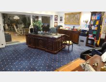 Elegantes Büro mit dunklem, kunstvoll geschnitztem Holzschreibtisch und Ledersessel auf einem blauen Teppich mit Muster. Im Hintergrund befindet sich eine Holzkommode, ein Bücherregal, verschiedene Wanddekorationen sowie ein großer Glaseinsatz, der zu einem Wintergarten mit Sitzmöbeln führt.