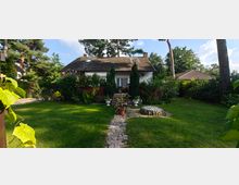 Einfamilienhaus mit Satteldach inmitten eines gepflegten Gartens, umgeben von Bäumen und Sträuchern; im Vordergrund ein gepflasterter Weg, ein kleiner Holzbrunnen und grüne Rasenflächen.