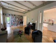 Wohnzimmer mit hellen Holzdielen, einer großen Bücherwand, einem schwarzen Klavier und modernen Möbeln. Rechts führt eine Tür in eine offene, weiße Küche, und links öffnet sich eine Glastür zu einer Terrasse mit Gartenblick.