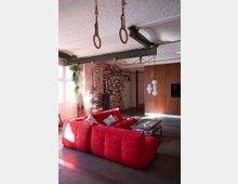 Ein Wohnraum mit industriellem Stil, gekennzeichnet durch freiliegende Backsteinwände, sichtbare Metallträger und Betonboden. Im Vordergrund steht ein rotes, gepolstertes Sofa um einen Glastisch, während Bücherstapel, Hängepflanzen und Turnringe an der Decke weitere dekorative Details bieten.