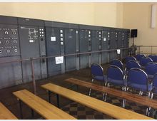 Ein Raum mit alten, grauen Schalttafeln an der Wand, davor Metallgeländer und ein Boden mit Schachbrettmuster. Im Vordergrund stehen einfache Holzsitzbänke und Stuhlreihen mit blauen Polstern.