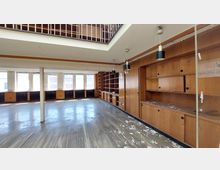 Ein leerer Raum mit einer Wand aus Holzregalen und Schränken, großen Fenstern, die viel Tageslicht hereinlassen, und einem Boden aus abgenutztem Holz. Die Decke verfügt über eine offene Galerie und herabhängende Lampen, während im Raum verstreut Schutt sichtbar ist.