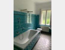 Badezimmer mit türkisfarbenen Fliesen, einer weißen Badewanne mit Handbrause, einem Waschbecken, einem WC und einem Fenster mit blickdichten Scheiben, das Tageslicht hereinlässt. Der Boden ist mit quadratischen, grauen und weißen Fliesen ausgelegt.