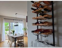 Moderner Essbereich mit Holzfußboden, Esstisch und Stühlen vor großen Schiebetüren mit weißen Vorhängen, die auf eine Terrasse und einen Garten führen. An der Wand im Vordergrund hängt ein schwarzes Metall-Weinregal mit Holzablagen für Flaschen und hängenden Weingläsern.