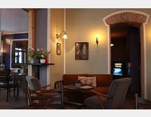 Gemütlicher Innenraum eines Cafés mit vintage-inspirierter Einrichtung. Zu sehen sind eine kleine Sitzecke mit Sofa, Holzstühlen, rundem Tisch, Wandleuchten, gemalten Bildern an einer pastellfarbenen Wand sowie ein gemauerter Rundbogen, der in einen weiteren Bereich führt.