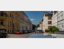 Straßenszene in einer städtischen Wohngegend mit mehrstöckigen Wohnhäusern auf beiden Seiten, teils in hellen Farben und mit klassischen Fassadenelementen. Geparkte Autos säumen die Kopfsteinpflasterstraße, und im Hintergrund sind Bäume und der blaue Himmel zu sehen.