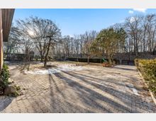 Großzügiger, gepflasterter Hof mit vereinzelten Schneeresten und langen Schatten, umgeben von Hecken und kahlen Bäumen. Im Hintergrund liegt ein bewaldeter Hang, rechts steht eine kleine Holzkonstruktion mit Rutsche.