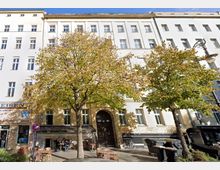 Fassade eines mehrstöckigen Wohngebäudes mit heller Putzfassade und großen Fenstern, flankiert von zwei breiten Bäumen mit herbstlich gefärbtem Laub. Im Erdgeschoss befinden sich ein Reisebüro und ein Café mit Sitzgelegenheiten im Außenbereich.