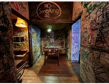 Schmaler Innenraum eines alternativen Bars oder Clubflurs mit Holzboden, dessen Wände und Decke vollständig mit Graffiti und Schriftzügen bedeckt sind. In der Mitte steht ein Tischkicker unter einer Deckenlampe; links hängt eine Stehlampe, oben ist ein „No Smoking“-Hinweis auf die Decke gemalt.