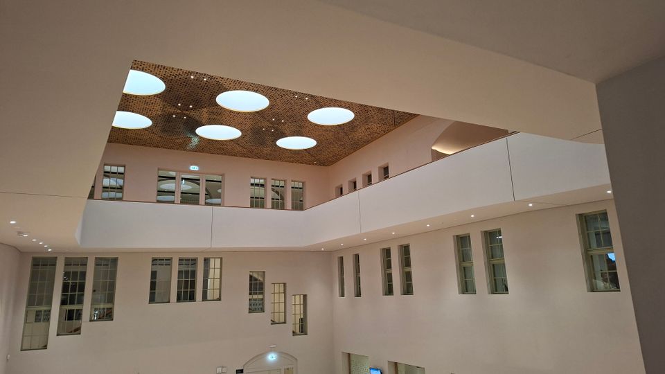Innenansicht eines modernen, mehrstöckigen Atriums mit umlaufenden Galerien, weißen Wänden und vielen rechteckigen Fenstern. In der hohen Decke sind mehrere große, runde Oberlichter eingelassen, ergänzt durch eingelassene Spotbeleuchtung entlang der Galerie.