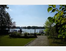 Gartenbereich mit einem Kiesweg, der zu einem Wasserufer führt; im Hintergrund befindet sich ein See mit Booten, umgeben von Bäumen und dichter Vegetation.