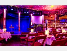 Innenansicht eines Varietés oder Theaters mit einer Bühne im Hintergrund, umgeben von roten Samtsäulen und blauer Beleuchtung. Im Vordergrund befinden sich Tische mit weißen Tischdecken und bestuhlte Sitzreihen, die von warmem Kerzenlicht beleuchtet werden.