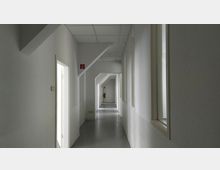 Langer, schmaler und weiß gestrichener Flur mit glatten Wänden und abgehängter Rasterdecke. Es gibt mehrere Türen und Fenster an den Seiten, ein Feuerlöscher ist am Ende des Flurs sichtbar.