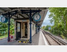 Bahnhofplattform mit überdachtem Wartebereich, einer Aufsichts-Kabine und großen runden Bahnhofuhren an verzierten Metallstützen. Rechts befinden sich die Gleise, umgeben von Bäumen und einem Grünstreifen.