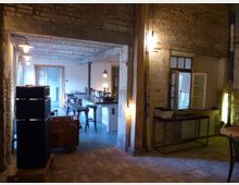 Ein rustikaler Innenraum mit freiliegenden unverputzten Backsteinwänden und Holzbalken. Im Vordergrund steht ein Tisch, im Hintergrund ist eine modern ausgestattete Küchenzeile mit Barhockern, auf einem Boden aus Holzdielen und Ziegelsteinfliesen.