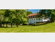 Ein weißes Gebäude mit rotem Ziegeldach und mehreren Fenstern, umgeben von grünen Bäumen und einer Rasenfläche in einer Parklandschaft. Links im Hintergrund ist eine kleine Steinbrücke sichtbar.