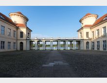 Schlosshof eines historischen Gebäudes mit zwei symmetrischen Flügeln und roten Ziegeldächern, verbunden durch einen Säulengang. Im Hintergrund sind ein See und Bäume unter klarem Himmel zu sehen.
