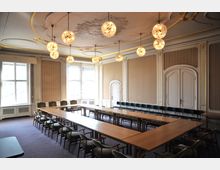 Eleganter Tagungsraum mit langen, U-förmig angeordneten Holztischen und gepolsterten Stühlen. Die Wände sind mit goldenen Verzierungen und Stofftapeten dekoriert, die Decke ist mit Stuckornamenten und mehreren kugelförmigen, leuchtenden Lampen gestaltet.
