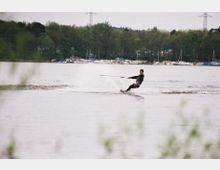 Eine Person fährt Wasserski auf einem breiten See, umgeben von einem bewaldeten Ufer mit zahlreichen geparkten Booten im Hintergrund. Der Himmel ist grau und es sind Strommasten in der Ferne sichtbar.