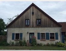 Einfamilienhaus mit Satteldach, verputztem Erdgeschoss und einer mit Holz verkleideten oberen Fassade. Das Haus hat Fenster mit blauen Fensterläden, eine zentrale Tür und einen kleinen Garten mit Blumen vor der Fassade.
