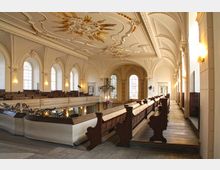 Innenraum einer historischen Kirche mit sichtbaren Emporen und Holz-Bänken. Die weiße Decke ist mit goldenen Verzierungen geschmückt, große Rundbogenfenster lassen Licht herein und Wandleuchten erhellen den Raum.