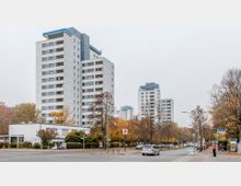 Mehrstöckige Wohnhochhäuser mit weißer Fassade und blauen Akzentdächern, umgeben von Herbstbäumen und einer Straßenkreuzung mit Autos und Fußgängern. Im Vordergrund ein flaches Gebäude, das als Ärztehaus gekennzeichnet ist.