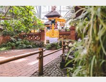 Ein kleiner balinesischer Tempel mit traditionellen Strohdächern und gelben Stoffverzierungen steht inmitten eines tropischen Gartens mit üppigen Pflanzen, Ziegelmauern und einem gepflasterten Weg. Die Anlage ist durch Bambusgeländer abgegrenzt, umgeben von exotischen Blumen und Grün.